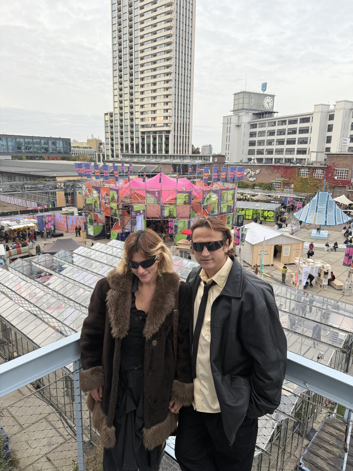 Mahir and Katya on a walkway above Strijp-S during Dutch Design Week, with the green Trudo tower behind them